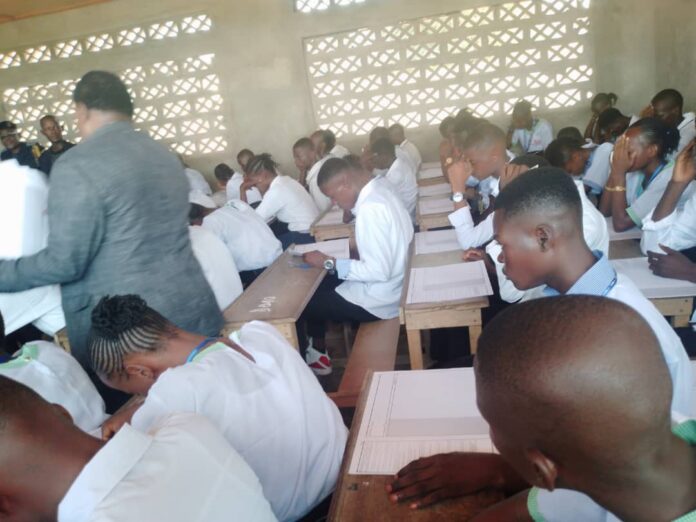 Salle d'examen à Bandundu avec des pupitres, des copies et une évacuation d'urgence vers un hôpital en arrière-plan