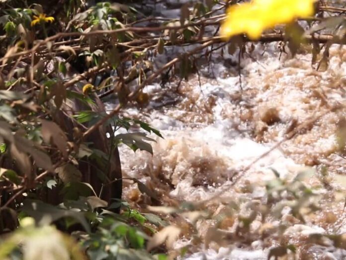 Route poussiéreuse blanchâtre à Kwampisha avec une conduite déversant des eaux usées mousseuses dans une rivière, pollution par les