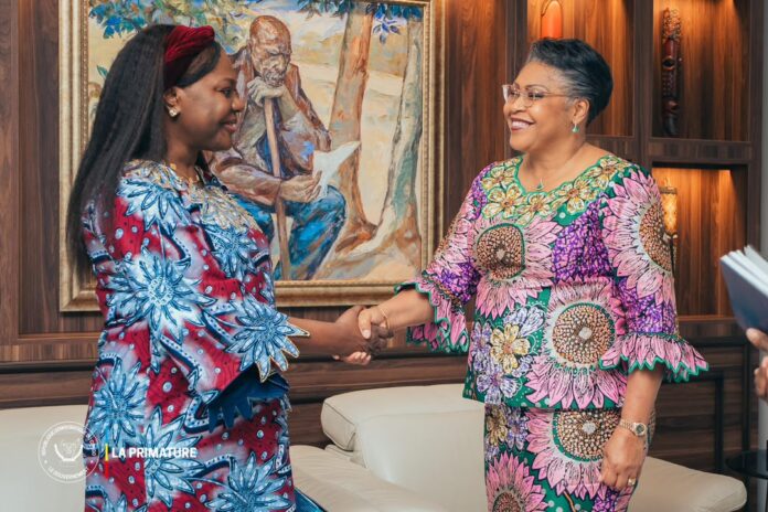 Première Ministre Judith Suminwa serrant la main de Brigitte Mukanga dans un salon officiel avec drapeaux RDC et ONU