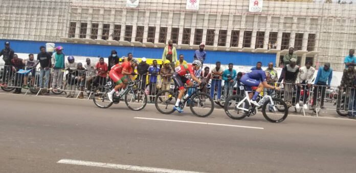 Cycliste en plein effort sur route, avec d'autres coureurs et un paysage camerounais