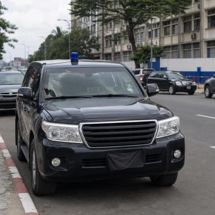 Véhicule noir avec gyrophare bleu sur le toit et plaque d'immatriculation recouverte d'un tissu, circulant dans une rue de Kinshasa