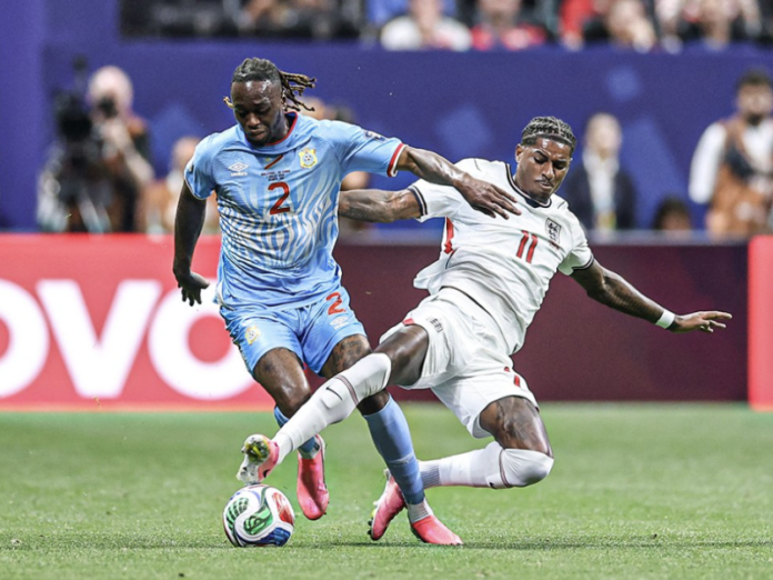 Joueur de football congolais en maillot jaune défendant contre un attaquant anglais lors d'un match de Coupe du Monde