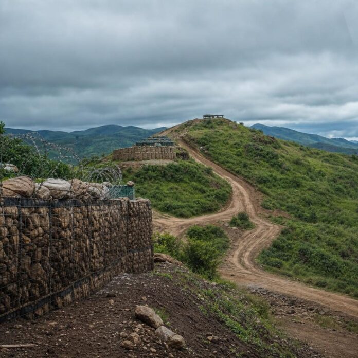 Paysage de l'est congolais avec collines, route strategique, positions militaires lointaines et atmosphere de tension