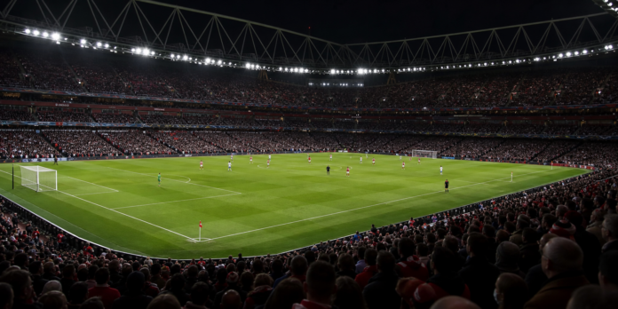 Vue large d un grand stade londonien avec une equipe rouge et blanche face a une equipe claire.