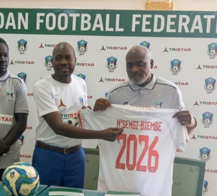 Entraîneur vu de dos sur le bord d'un terrain de football, joueurs en action au loin, ambiance de match international