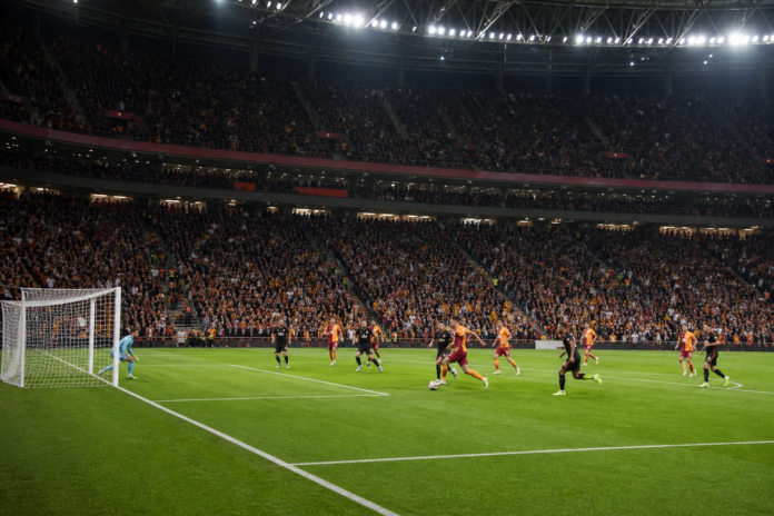 Illustration editoriale d un match de football sous les projecteurs avec une equipe jaune et rouge face a une equipe sombre.