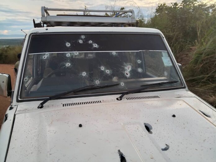 Un Land Cruiser blanc avec impacts de balles et vitres brisées sur une piste poussiéreuse, dans un paysage de brousse minière