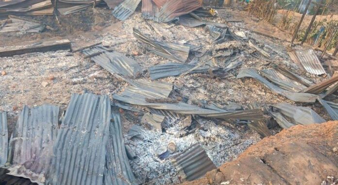 Deux maisons en planches consumées par le feu, avec des débris calcinés et une moto brûlée au premier plan