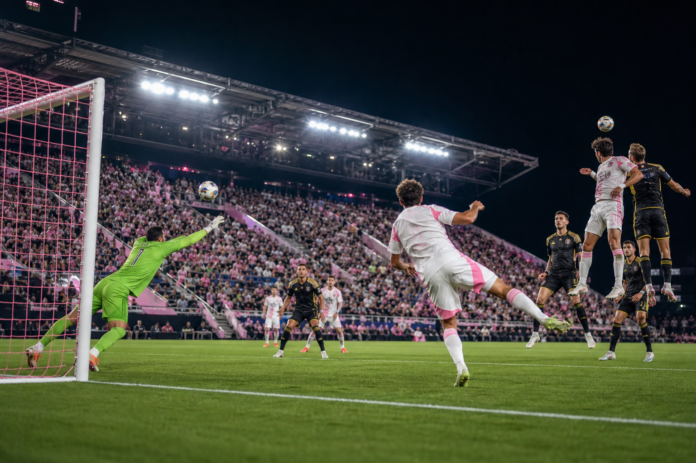 Scene de match nocturne dans un stade americain entre une equipe blanche a touches roses et une equipe sombre.