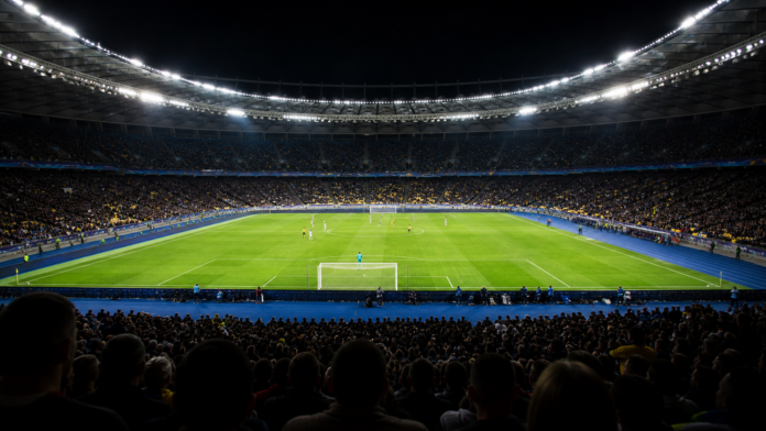 Stade de football europeen illumine avec joueurs lointains non identifiables et atmosphere de soiree, sans logo ni visage visible.