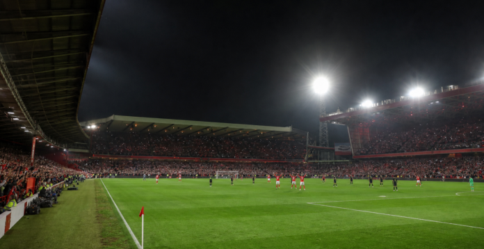 Vue large d un match sous les projecteurs avec une equipe rouge face a une equipe sombre.