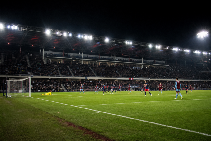 Vue large d un match europeen nocturne entre deux equipes aux couleurs sombres et bordeaux.