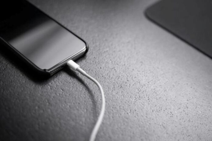 Un smartphone allumé posé sur une table de nuit, son câble de recharge branché, dans une chambre sombre