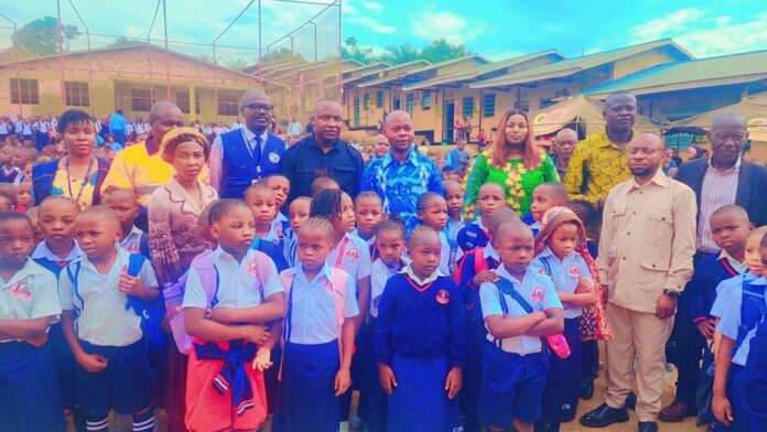 Élèves en uniforme alignés de dos dans la cour d'une école à Durba, avec un podium et des seaux d'eau pour le lavage des mains