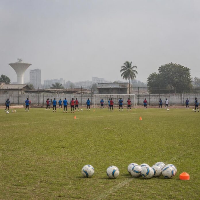 Un joueur de football en tenue d'entraînement bleu et rouge s'exerce sur un terrain à Kinshasa, entouré de cônes