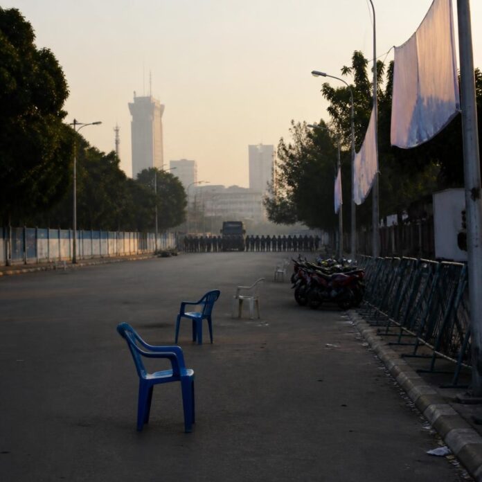 Rue de Kinshasa bordée de vendeurs et de barrières métalliques avant une marche de l'opposition, avec agents en tenue antiémeute au loin
