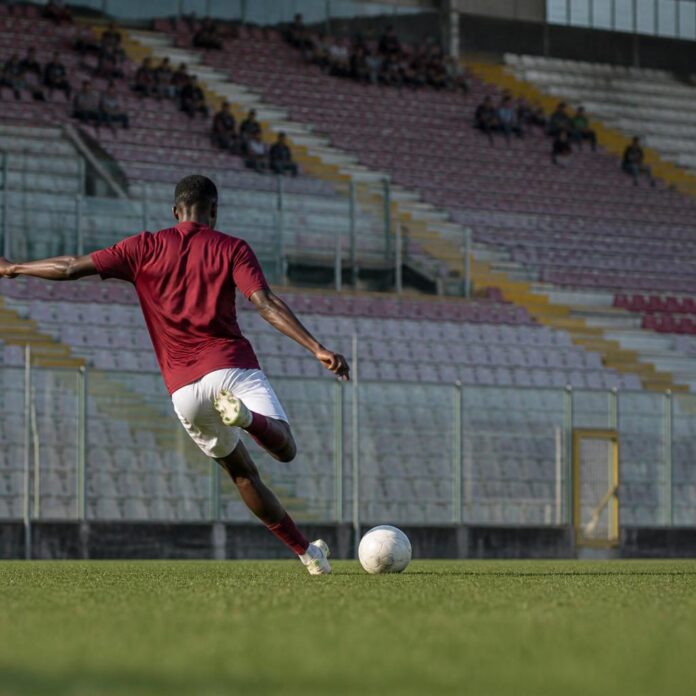 Joueur de football en maillot amarante s'apprêtant à frapper un ballon lors d'un match dans un stade italien, vu de dos