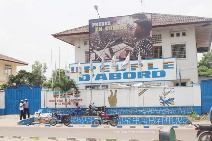 Salle de réunion à Kinshasa avec micros, documents et drapeaux évoquant une déclaration politique de l’UDPS