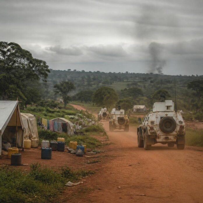 Véhicules blindés onusiens et paysage rural de l’est de la RDC, avec route de terre et campement de déplacés