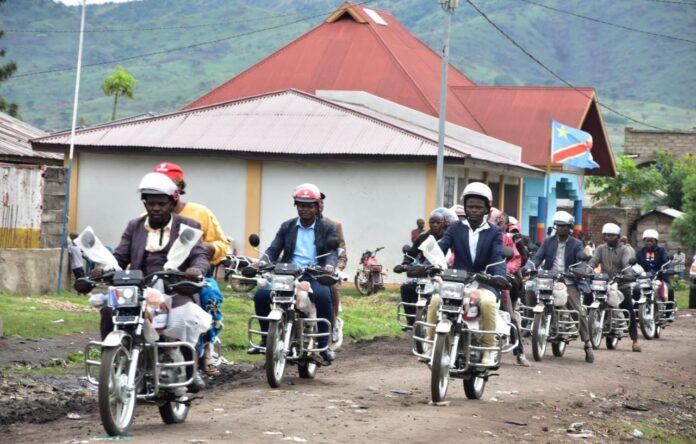 Groupe de personnes en moto avec casque sur une route devant des maisons avec un drapeau bleu rouge et jaune