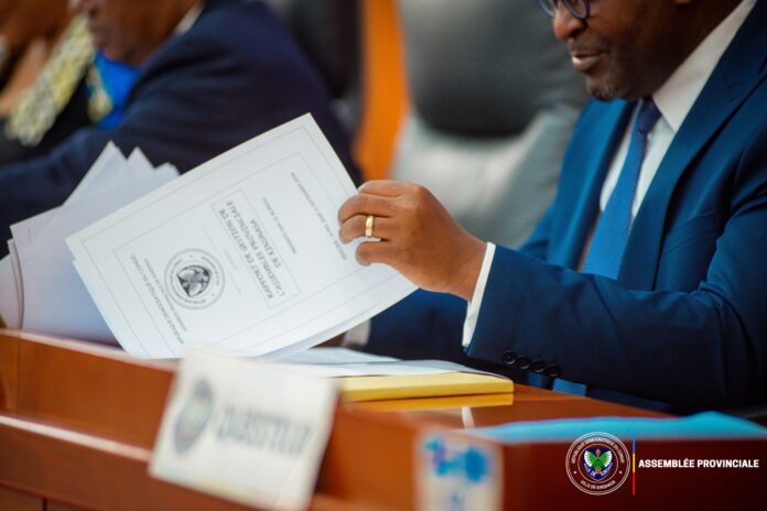 Hémicycle de l’Assemblée provinciale de Kinshasa avec pupitres, dossiers budgétaires et microphones, sans visage reconnaissable