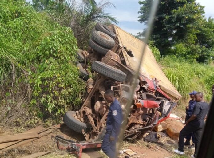 Camion poids lourd renversé au bord d'une route dégradée, bidons d'huile de palme et marchandises éparpillés, sans personne visible
