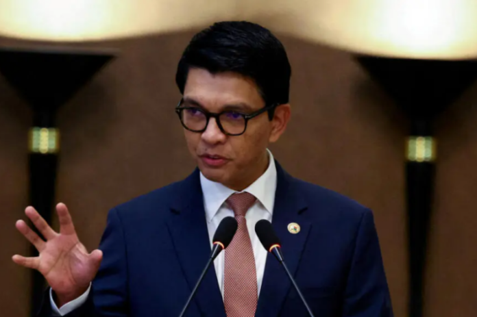 Homme avec lunettes, costume bleu et cravate rose parlant devant deux micros avec un fond flou