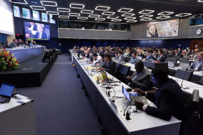 Salle de conférence internationale avec pupitres, micros, écrans et documents sur les télécommunications, ambiance de négociations