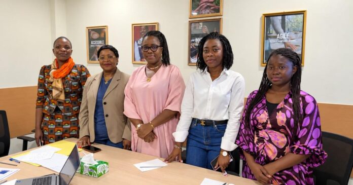 cinq femmes debout dans une salle de réunion avec des cadres au mur et une table avec documents et ordinateur
